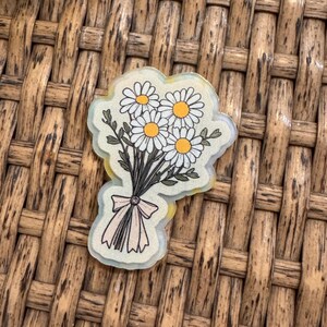May include: A clear sticker featuring a bouquet of daisies. The daisies have white petals and yellow centers. The bouquet is tied with a light pink ribbon. The sticker is on a woven, brown background.