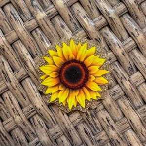 May include: A circular sunflower emblem featuring vibrant yellow and orange petals surrounding a dark brown center. The sunflower is set against a scalloped-edge background covered in a fine, shimmering gold glitter finish.