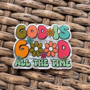 May include: A colorful, retro-style pin with the words "GOD IS GOOD ALL THE TIME" in bubble letters. The pin features orange, pink, green, and teal colors, with flower accents. The pin is set against a textured, woven background.