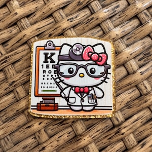 May include: A square pin featuring a cartoon cat dressed as an eye doctor, wearing a white lab coat, red bowtie, and black glasses. The cat holds a pointer next to an orange eye chart with black letters. The pin has a gold glitter border.