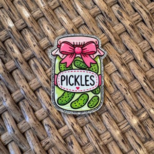May include: A glittery jar-shaped pin featuring a pink bow and the word "PICKLES" on a white oval. The jar is filled with green pickles and has a silver outline. The pin is set against a woven brown background.