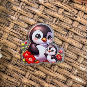 May include: A decorative sticker featuring a mother penguin and a baby penguin. The penguins are black and white with pink cheeks and orange beaks. Red flowers and green leaves surround the penguins, with a silver glitter border.