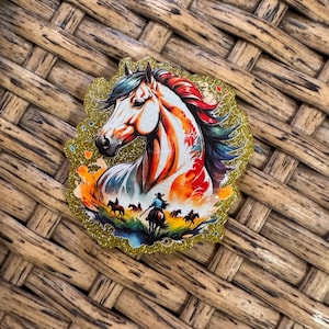 May include: A colorful horse-themed sticker with a gold glitter border. The horse has a white face, a red and orange mane, and a blue and green tail. The sticker also features a cowboy riding a horse with other horses in the background.