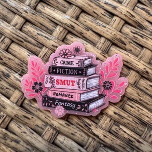 May include: A pink and black sticker featuring a stack of books with the titles "Crime", "Fiction", "Smut", "Romance", and "Fantasy". The sticker has a floral design and glitter accents.