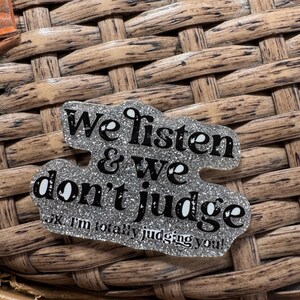 May include: A glittery silver pin with the words "We listen & we don't judge" in black lettering. Below the main text is the phrase "JK I'm totally judging you!" in a smaller font. The pin is on a woven surface.