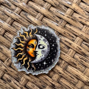 May include: A round, glitter-edged pin featuring a celestial design. The image is split between a sun with orange and yellow hues and a crescent moon with white and gray tones. The background is black with white stars.
