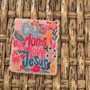 May include: A decorative, glittery square with the words "God Moms Love Jesus" in blue, orange, and pink. The design is surrounded by floral illustrations in shades of pink, orange, and red.