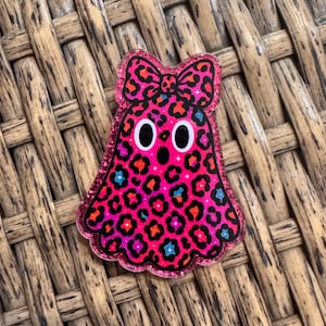 May include: A ghost-shaped acrylic pin featuring a vibrant hot pink base with a black leopard print pattern. The ghost has two large white oval eyes with black pupils, a small black circular mouth, and a bow on top. The edges have a glitter finish.