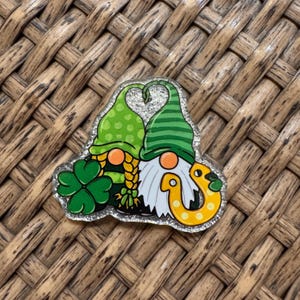 May include: A St. Patrick's Day themed pin featuring two gnomes with green hats and orange noses. One gnome has a green shamrock, and the other has a yellow horseshoe. The pin has a silver glitter border.
