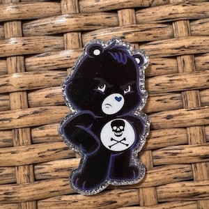 May include: A black Care Bear-style pin with a skull and crossbones on its belly. The bear has an angry expression, a blue heart on its chest, and a glittery silver border. The pin is set against a woven background.