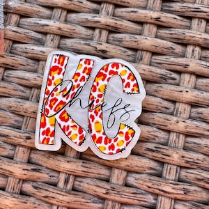 May include: A clear, decorative sticker with the letters "KC" in red, yellow, and white leopard print. The word "Chiefs" is written in black cursive script across the letters. The sticker is on a woven brown surface.
