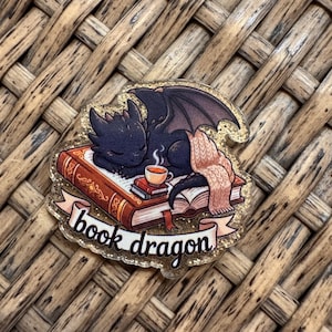 May include: A glittery acrylic pin featuring a black dragon sleeping on a stack of books with a small cup of tea and a pink knit blanket. A banner at the bottom displays the text book dragon in black script against a light pink background.
