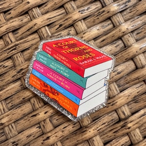 May include: A glittery acrylic pin featuring a stack of five books from the A Court of Thorns and Roses series by Sarah J. Maas. The spines are colored red, teal, pink, blue, and orange, each displaying the title and author name in clear text.