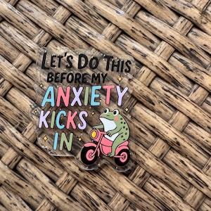 May include: A clear acrylic pin with the text "Let's Do This Before My Anxiety Kicks In." The pin features a cartoon frog riding a pink motorcycle. The text is in black, pink, blue, and purple.