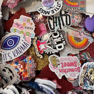 May include: A collection of colorful, glittery resin pins and badges with various designs and text. The pins feature phrases like "It's Karma," "Little Miss," and "Negative Thoughts," along with images of a rainbow, a guitar, and a baseball.