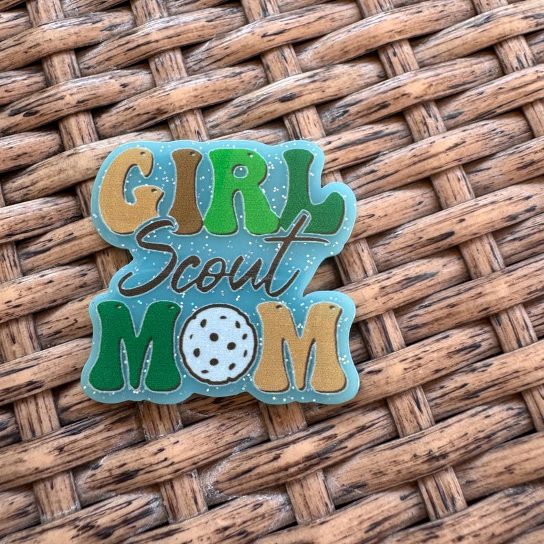 May include: A teal-colored pin with the words "GIRL Scout MOM" in a retro font. The word "GIRL" is in brown and green, "Scout" is in a script font, and "MOM" is in green and gold. A white and blue ball is in the center.