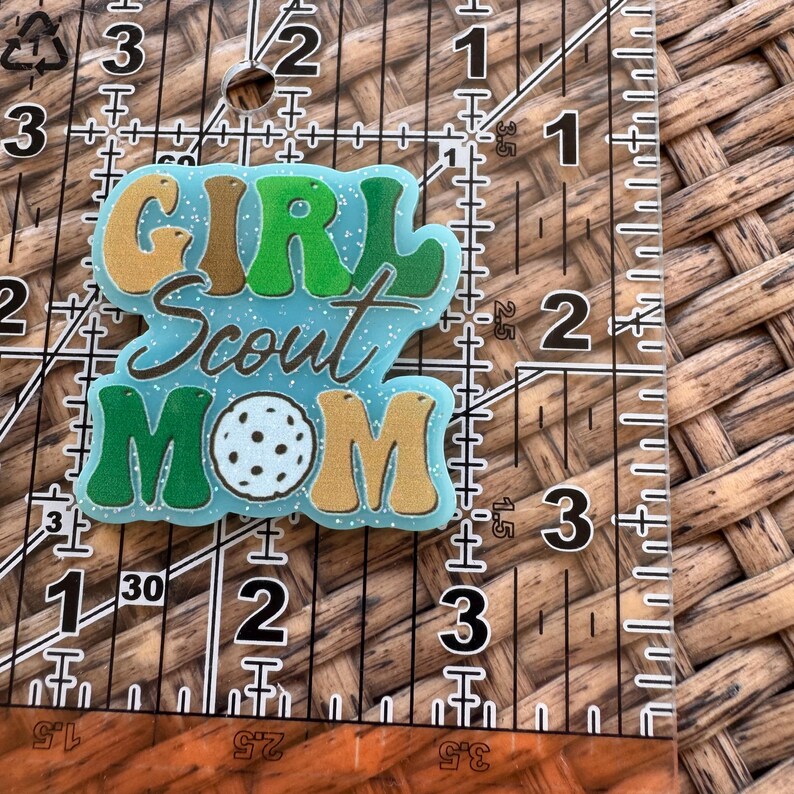 May include: A light blue Girl Scout Mom badge with glitter accents. The words "GIRL Scout MOM" are in green, gold, and white. A white and black pickleball is in the center of the word "MOM".