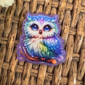 May include: A colorful owl sticker with a purple border. The owl has large blue eyes and is perched on a brown branch. The owl's feathers are a mix of blue, green, purple, and white. The background is a woven pattern.