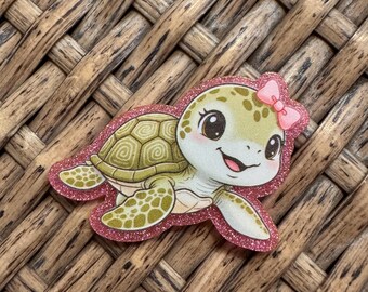 May include: A flat, die-cut sticker featuring a cartoon sea turtle with a light green shell and skin, large dark eyes, and a small pink bow on its head. The turtle is outlined by a shimmering, glittery pink border that follows its shape.