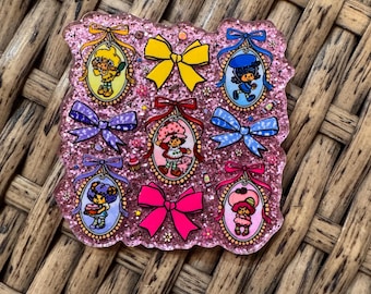 May include: A pink glittery pin featuring cartoon characters in oval frames, each topped with a colorful bow. The characters are in various poses, surrounded by small flowers and other decorative elements. The pin is on a woven surface.