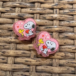 May include: Two heart-shaped Snoopy and Woodstock pins with a pink glitter border. Snoopy is holding a red heart, and Woodstock is next to him. The pins have a cartoon design and are set against a woven background.