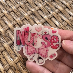 May include: A pink and white Nurse themed sticker with a syringe, stethoscope, and bow. The word "Nurse" is written in pink with heart and polka dot patterns. Small hearts and a nurse's hat are also included in the design.