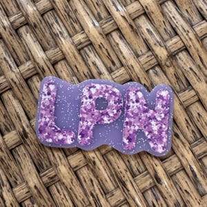 May include: A lavender-colored badge with the letters "LPN" in a floral pattern of purple and white. The badge has a glittery, speckled finish and is set against a woven, brown background.