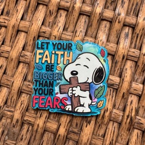 May include: A colorful pin featuring Snoopy from Peanuts, holding a wooden cross. The pin has the text "LET YOUR FAITH BE BIGGER THAN YOUR FEARS" in a mix of colors. The background is a light blue with leaf accents.
