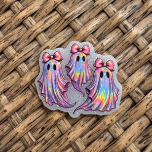 May include: A sticker featuring three ghosts with iridescent, holographic rainbow coloring and glitter accents. Each ghost has two black oval eyes and a pink bow on its head. The ghosts are arranged in a group on a translucent, glittery backing.