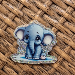 May include: A blue cartoon elephant with large eyes and a trunk, sitting on a light blue surface. The elephant is surrounded by a glittery, silver border. The background is a woven, brown surface.