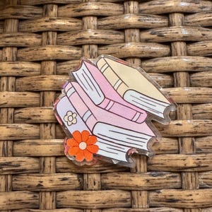 May include: A clear acrylic pin featuring a stack of books in shades of pink and beige. A small orange flower with a white center and a four-leaf clover design are also included in the design. The pin is set against a woven brown background.
