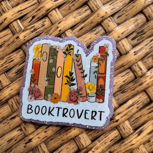 May include: A die-cut sticker featuring a row of colorful books in shades of orange, green, yellow, and blue, decorated with floral illustrations. The word BOOKTROVERT is printed in black capital letters at the bottom on a light blue background.