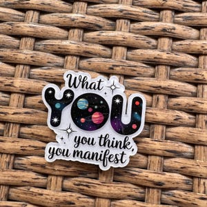 May include: A clear sticker with the words "What YOU you think, you manifest" in black script. The word "YOU" is designed with a space theme, featuring planets and stars in shades of blue, purple, and red.