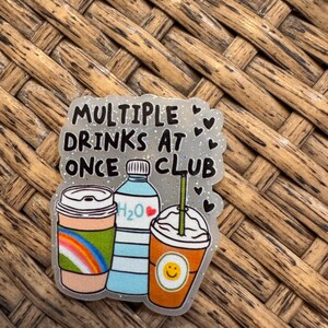 May include: A glittery sticker with the text "MULTIPLE DRINKS AT ONCE CLUB." The sticker features illustrations of a coffee cup with a rainbow, a water bottle, and a smoothie with a smiley face.