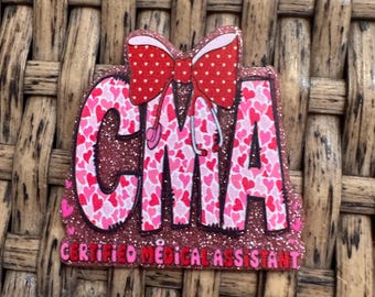 May include: A pink and red glittery CMA badge with a bow and hearts. The badge reads "CMA" in large letters, filled with pink hearts, and "CERTIFIED MEDICAL ASSISTANT" in smaller letters.