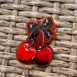 May include: A red glittery cherry pin with a black bow featuring a leopard print design. The pin has two cherries with a glossy finish and green stems. The bow is positioned above the cherries.
