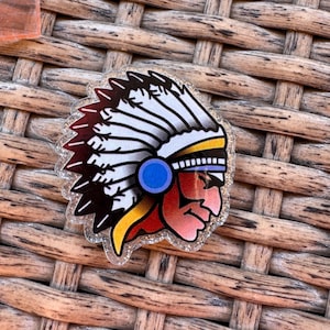 May include: A clear, glitter-edged pin featuring a Native American chief in profile. The design includes a feathered headdress with white and maroon feathers, a blue circle, and a red and yellow face.