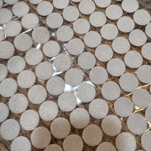 May include: Close-up of a collection of round, light gray hook and loop fasteners. The fasteners are arranged in a grid pattern, with a textured surface. The background is a light brown, possibly wood.