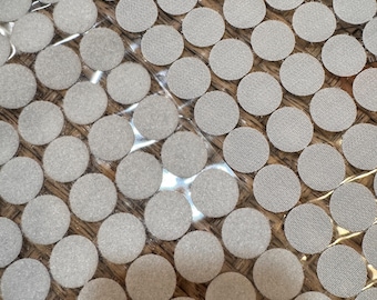 May include: Close-up of a collection of round, light gray hook and loop fasteners. The fasteners are arranged in a grid pattern, with a textured surface. The background is a light brown, possibly wood.