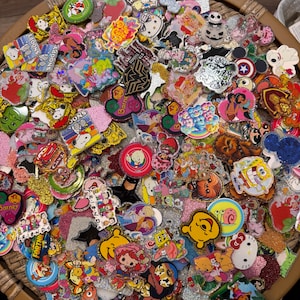 May include: A collection of colorful character pins, including Snoopy, Disney princesses, and SpongeBob. The pins feature glitter, enamel, and various shapes. The assortment includes a variety of pop culture and cartoon characters.