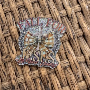 May include: A glittery, oval-shaped pin with the words "FALL FOR JESUS He Never Leaves" in pink and gold lettering. The pin features a cross and a bow with a plaid pattern. The background is silver glitter.