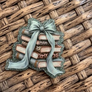 May include: A decorative sticker featuring a stack of four books tied together with a sage green ribbon. The books have alternating solid and leopard print covers in shades of brown, tan, and cream. The entire design is outlined in green glitter.