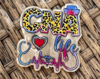 May include: A decorative acrylic badge featuring the text CNA life in a leopard print pattern. The design includes a red heart, a blue stethoscope forming a heartbeat line, a small caduceus symbol, and a blue nurse cap on the letter A.