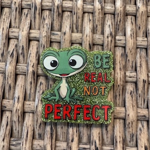 May include: A green frog-shaped pin with the words "BE REAL NOT PERFECT" in red and green. The pin has a glittery background and is set against a woven brown background.