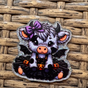 May include: A glittery acrylic charm featuring a cartoon cow with black and white fur, small black bat wings, and a purple bow. The cow wears a black skirt adorned with orange jack-o'-lanterns, set against a shimmering, holographic background.