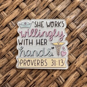 May include: A clear, glittery acrylic badge featuring a nurse cap icon and the text "SHE WORKS willingly WITH HER hands." alongside a caduceus symbol, a pill, and a heart. The bottom includes a yellow banner with the text "PROVERBS 31:13".