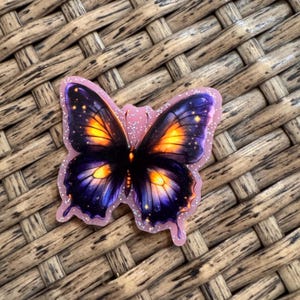 May include: A butterfly-shaped sticker featuring a dark purple and black wing pattern with glowing orange centers and small yellow star-like accents. The sticker has a light pink border with a subtle glittery finish throughout the design.