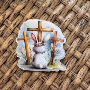 May include: A sticker featuring a gray bunny wearing a flower crown, seated before three wooden crosses. The bunny faces away, with long ears and a fluffy tail. Soft pastel colors and floral accents create a serene Easter theme.