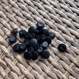 May include: A collection of small, black, round plastic fasteners with a flat top and a short post on the underside. The fasteners are scattered on a woven, light brown surface.