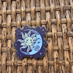 May include: A square-shaped sticker with rounded, cloud-like edges featuring a dark purple background with small white stars. The central design depicts a light blue crescent moon and a yellow sun with a face, both intertwined in a celestial pattern.